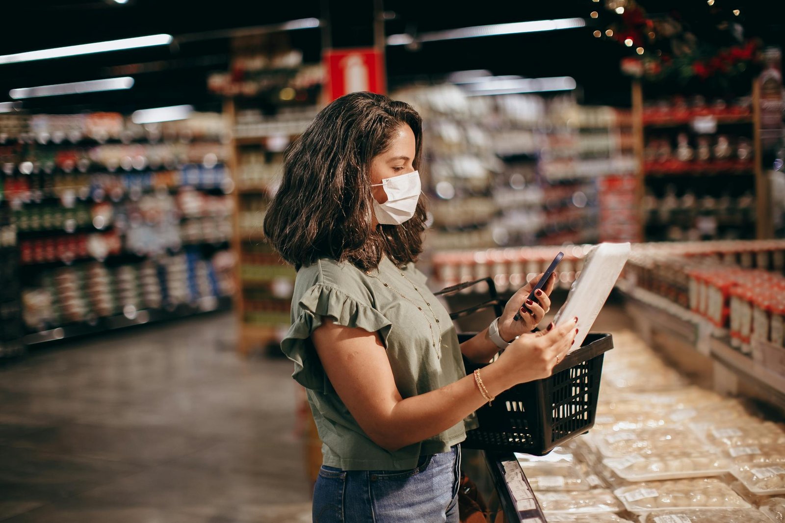 A woman shopping in a supermarket, wearing a face mask and using her phone to check products.
