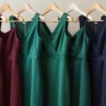 A collection of fall wedding guest dresses in rich jewel tones including burgundy, emerald, and navy arranged on elegant hangers against a neutral background