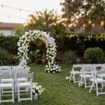 intimate small backyard wedding ceremony setup with white chairs and floral arch